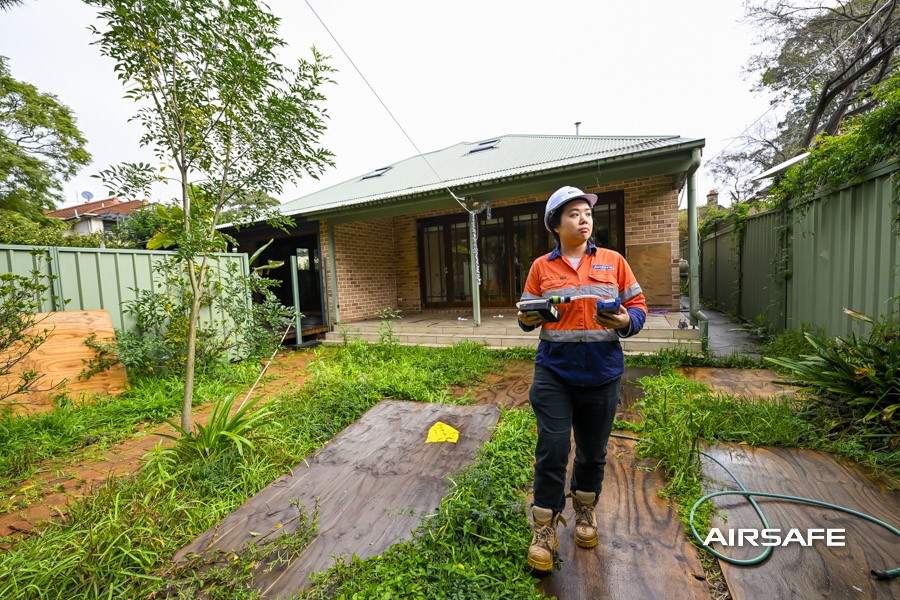 Airsafe consultant carrying air sampling equipment at the rear of an older brick home