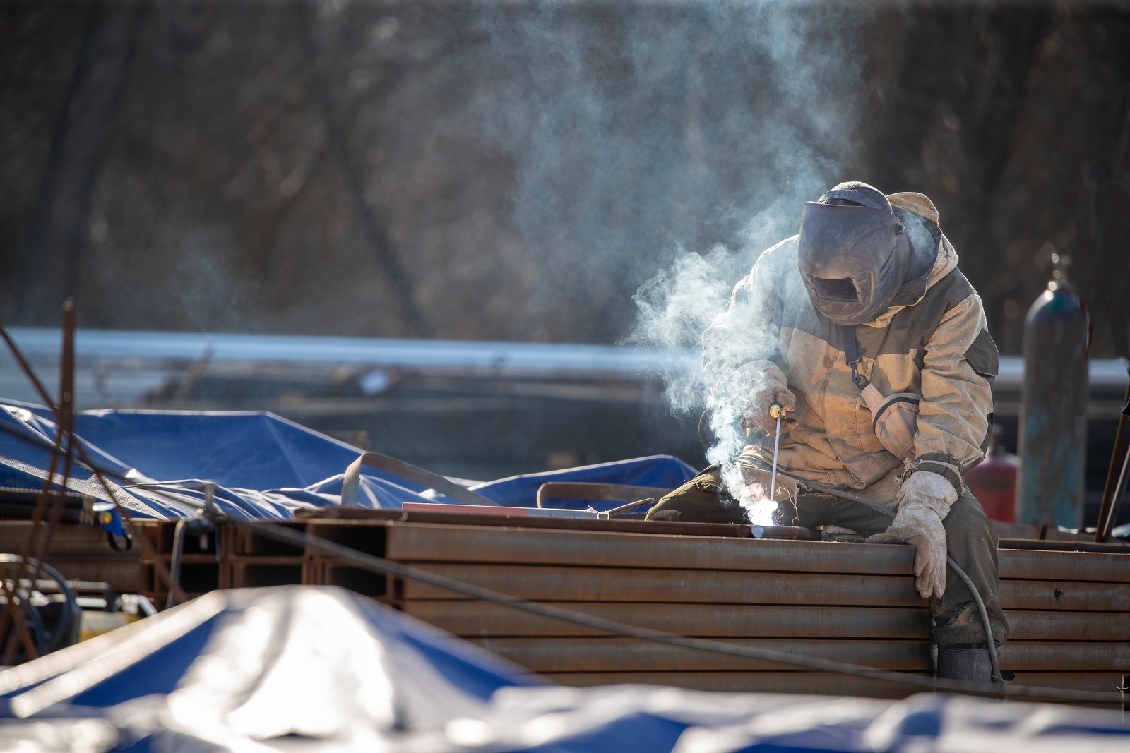 A welder works on stacked steel sections as welding fume rises above the arc