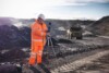 Person in orange hi-vis and a hard hat setting up a sound level meter on a tripod at an open-cut mine, with a haul truck behind