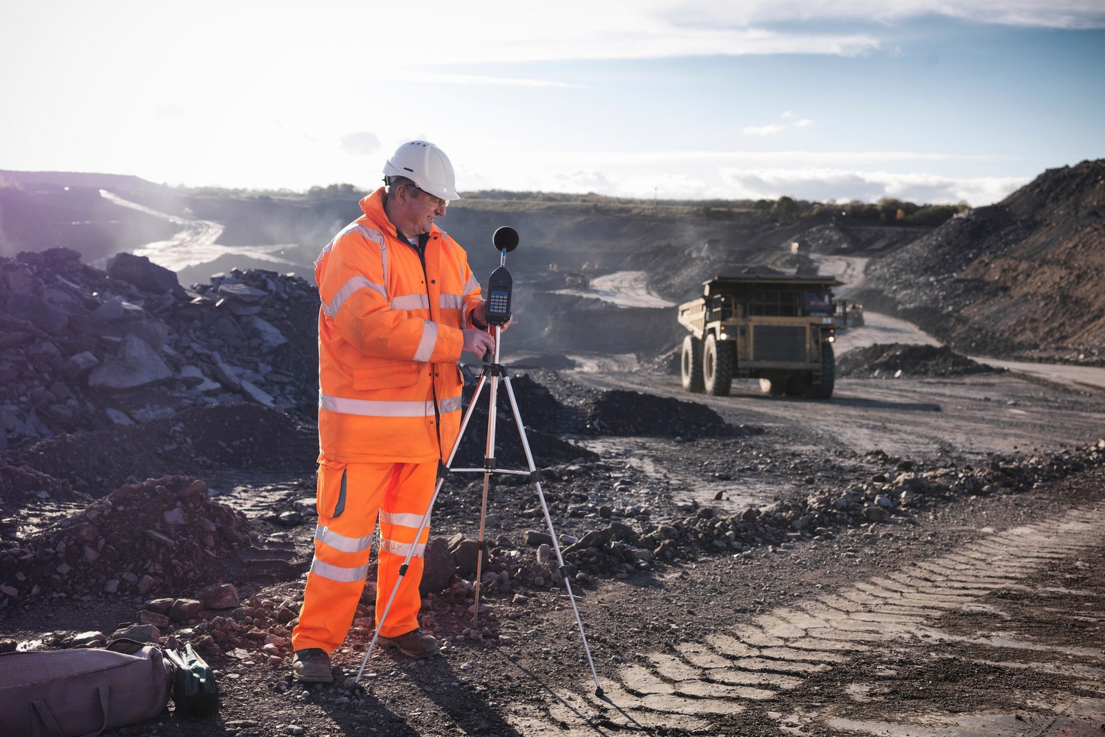 Person in orange hi-vis and a hard hat setting up a sound level meter on a tripod at an open-cut mine, with a haul truck behind